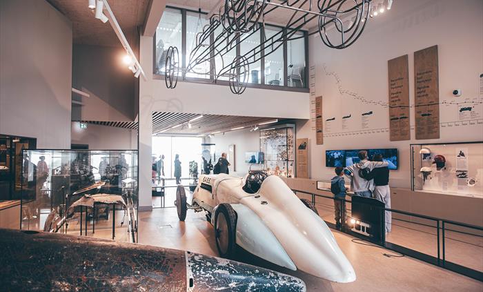 Wide angle of the main gallery interior of the museum. A large metal tail of the Bluebird car is in the foreground and the Babs car is in the middle g